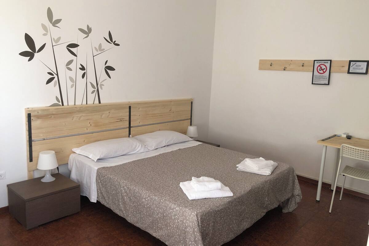 A neatly made bed with a grey floral bedspread, two white pillows, and a wooden headboard. There is a small bedside table with a lamp, a white chair, and a desk with a chair. A wall decoration of leaves is visible.