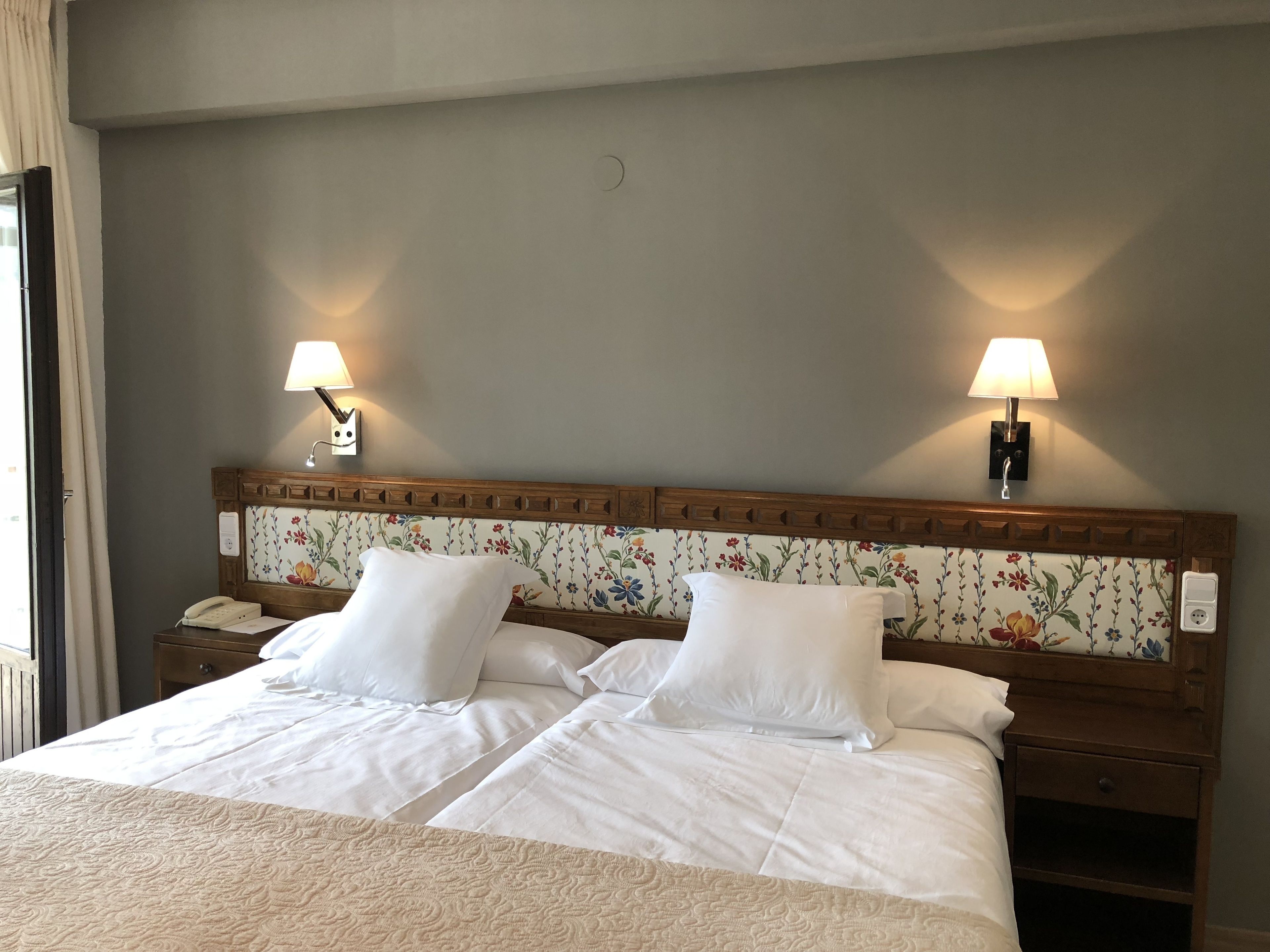 A double bed with white linens, two bedside tables, a floral headboard, and two wall-mounted lamps.