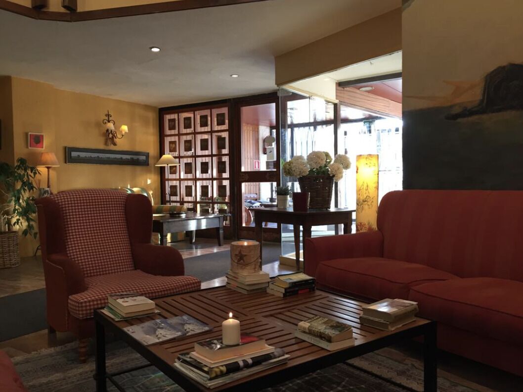 Un hall acogedor con un sillón estampado, un sofá rojo y una mesa de centro con libros y una vela. Se observa una estantería y un jarrón con flores al fondo.