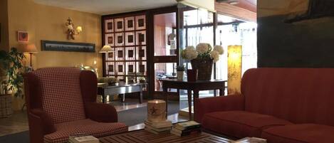 Un hall acogedor con un sillón estampado, un sofá rojo y una mesa de centro con libros y una vela. Se observa una estantería y un jarrón con flores al fondo.