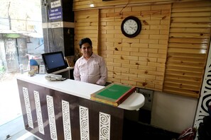 A reception desk with a clock, a computer, and a stack of books. - Hotel Suvidha Deluxe (New Delhi)