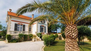 A two-story building with a red-tiled roof, a balcony with a stone balustrade, and a well-maintained garden with a palm tree. - 4 star villa with sea view, islands, natural area, private heated pool, spa (Port-Des-Barques)
