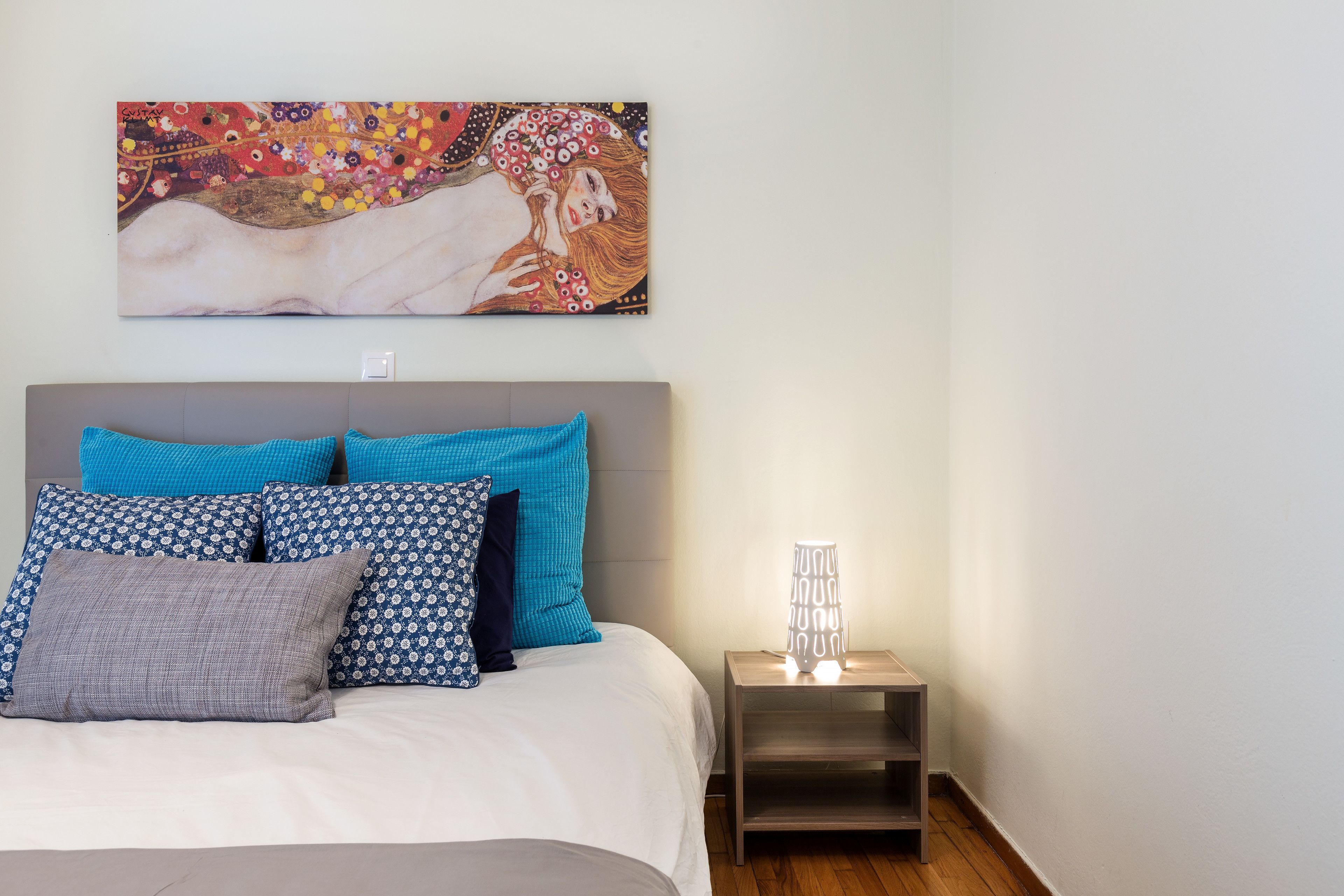 A bedroom with a bed, pillows, a nightstand, a lamp, and a wall painting.