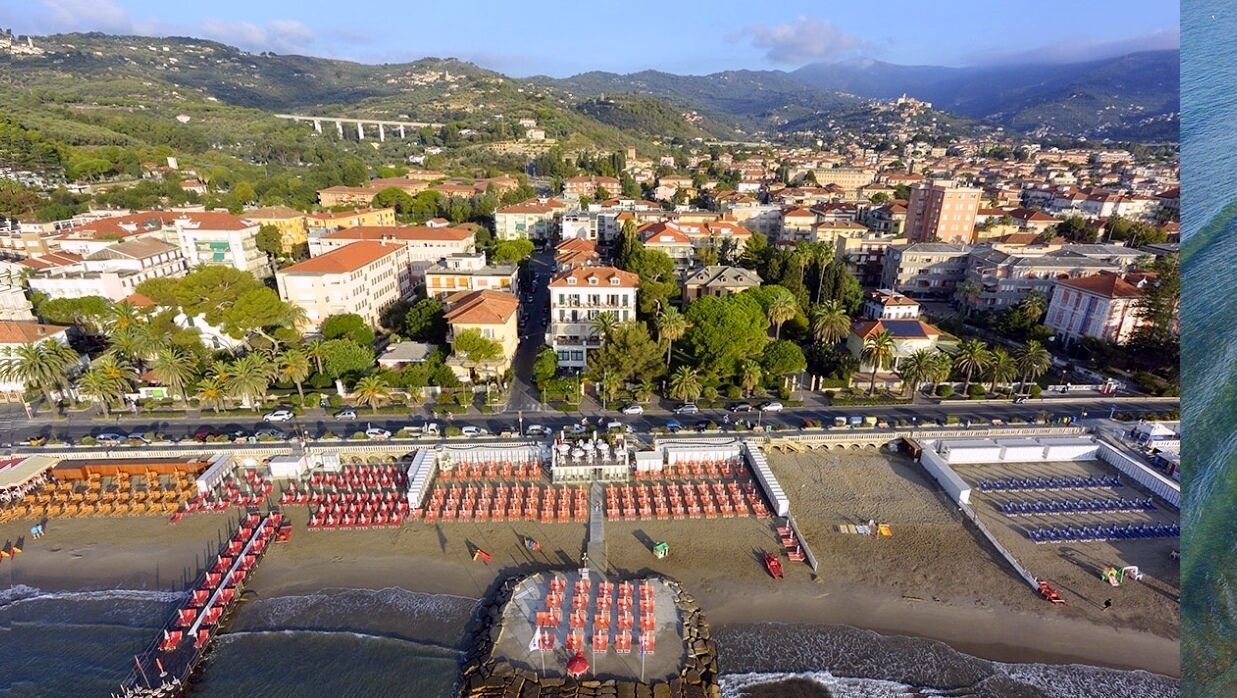 A coastal town with a beach area featuring rows of umbrellas and sun loungers, a promenade, and a backdrop of residential buildings and hills.