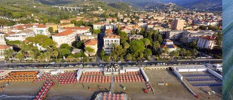 A coastal town with a beach area featuring rows of umbrellas and sun loungers, a promenade, and a backdrop of residential buildings and hills.