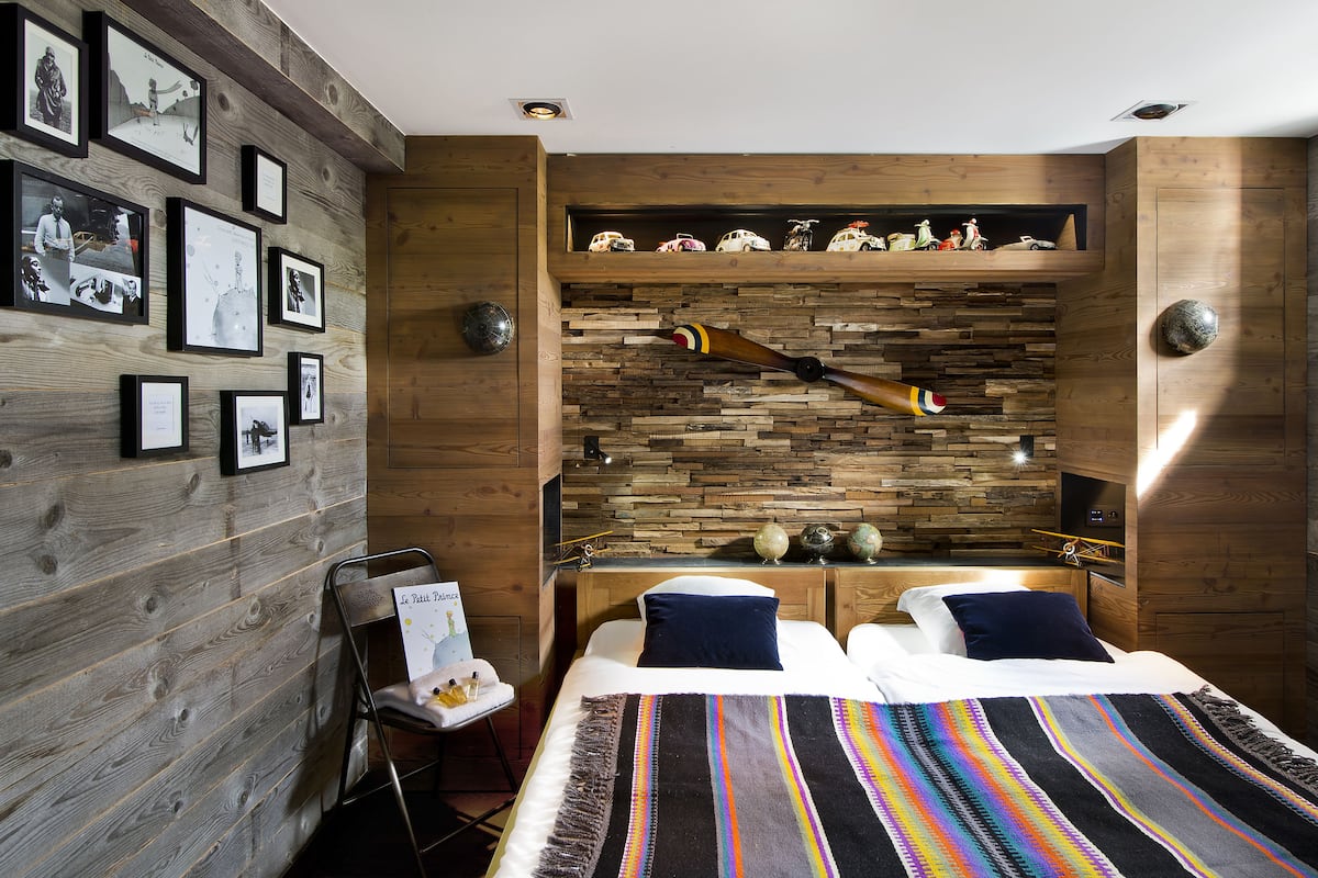 A modern hotel room with a wooden wall, a bed with a colorful striped blanket, a small desk with a globe and a toy airplane, and a shelf with model cars.