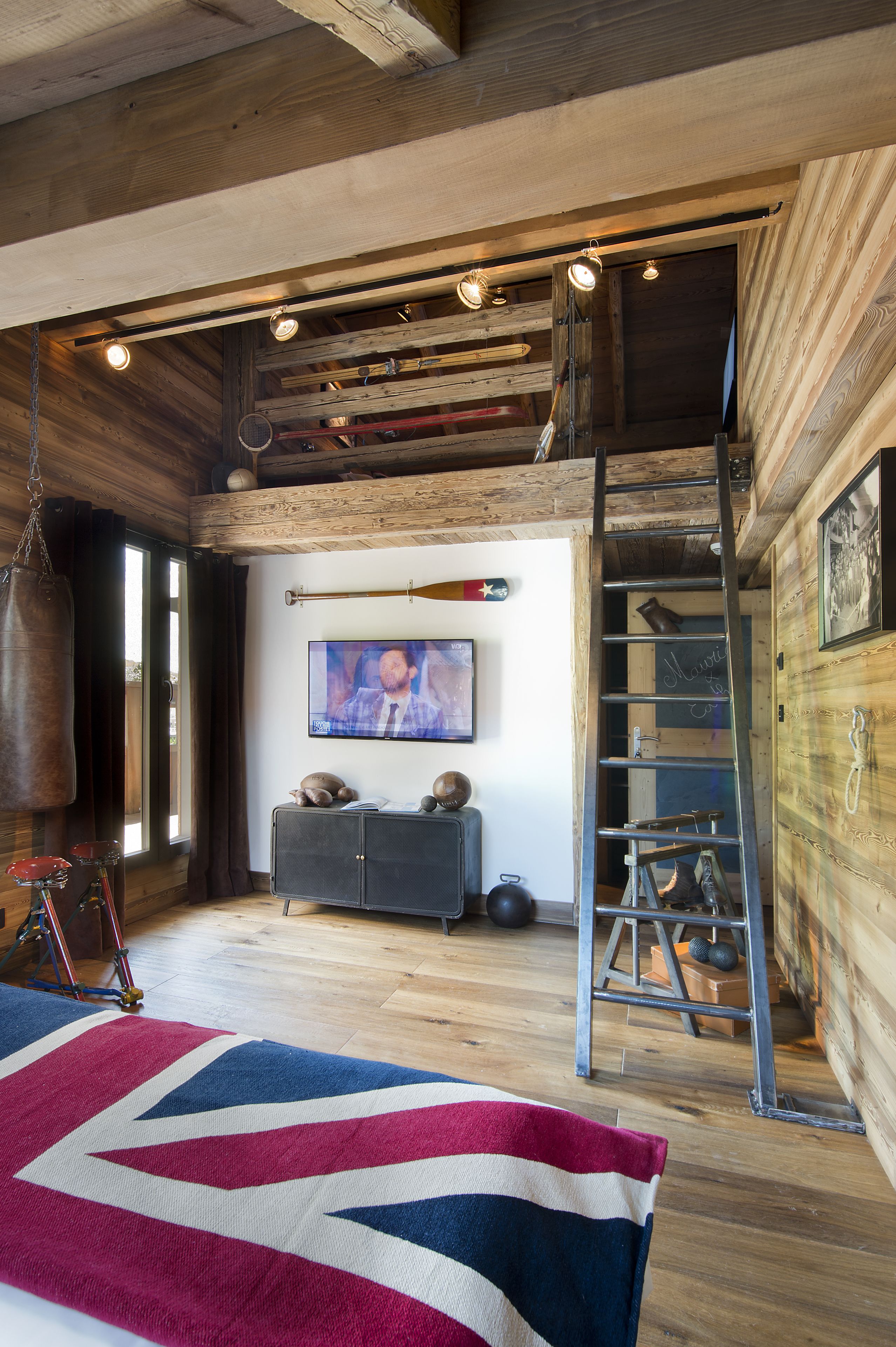 A spacious loft bedroom with a high ceiling, a large bed, a wooden ceiling with exposed beams, a television, a ladder, and a cozy fireplace.