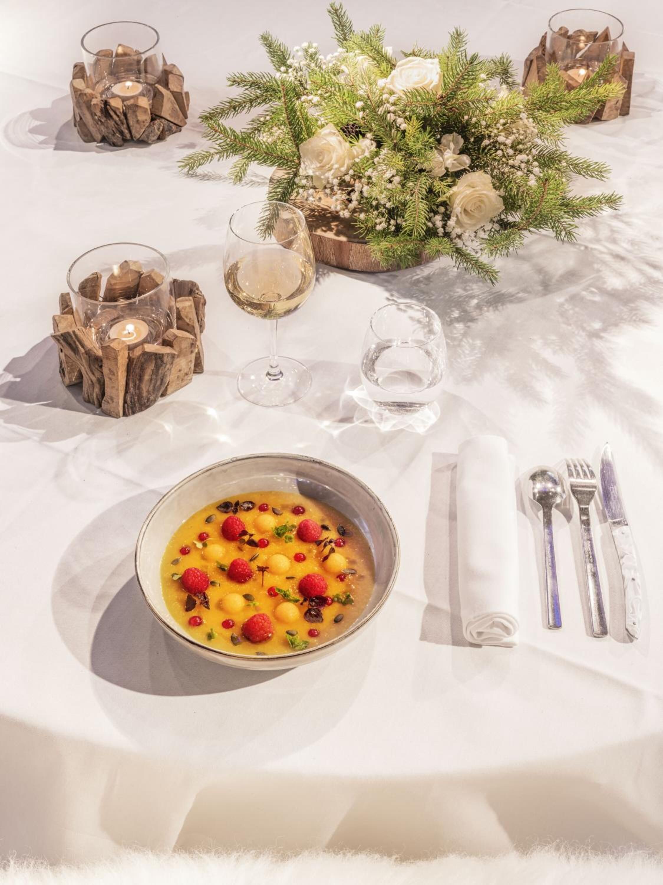 A table set with a bowl of soup garnished with raspberries and seeds, wine glasses, cutlery, and a floral centerpiece.