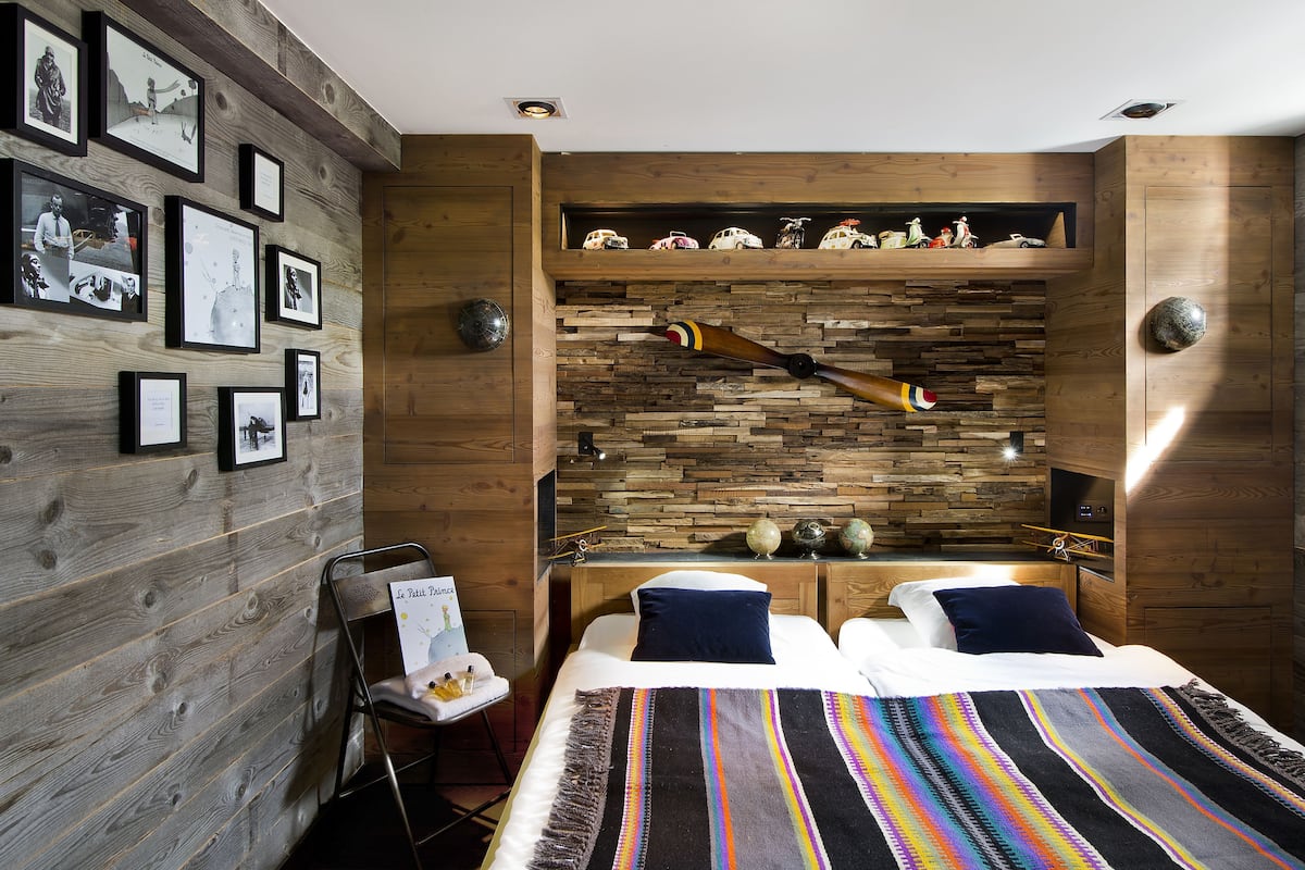 A bedroom with a wooden wall, a bed with a striped blanket, a small table with a globe, and a shelf with model cars.
