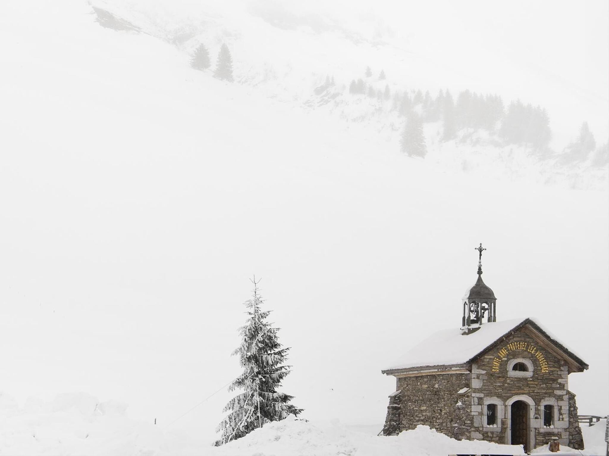 A stone church with a bell tower, surrounded by snow-covered mountains and a single evergreen tree.