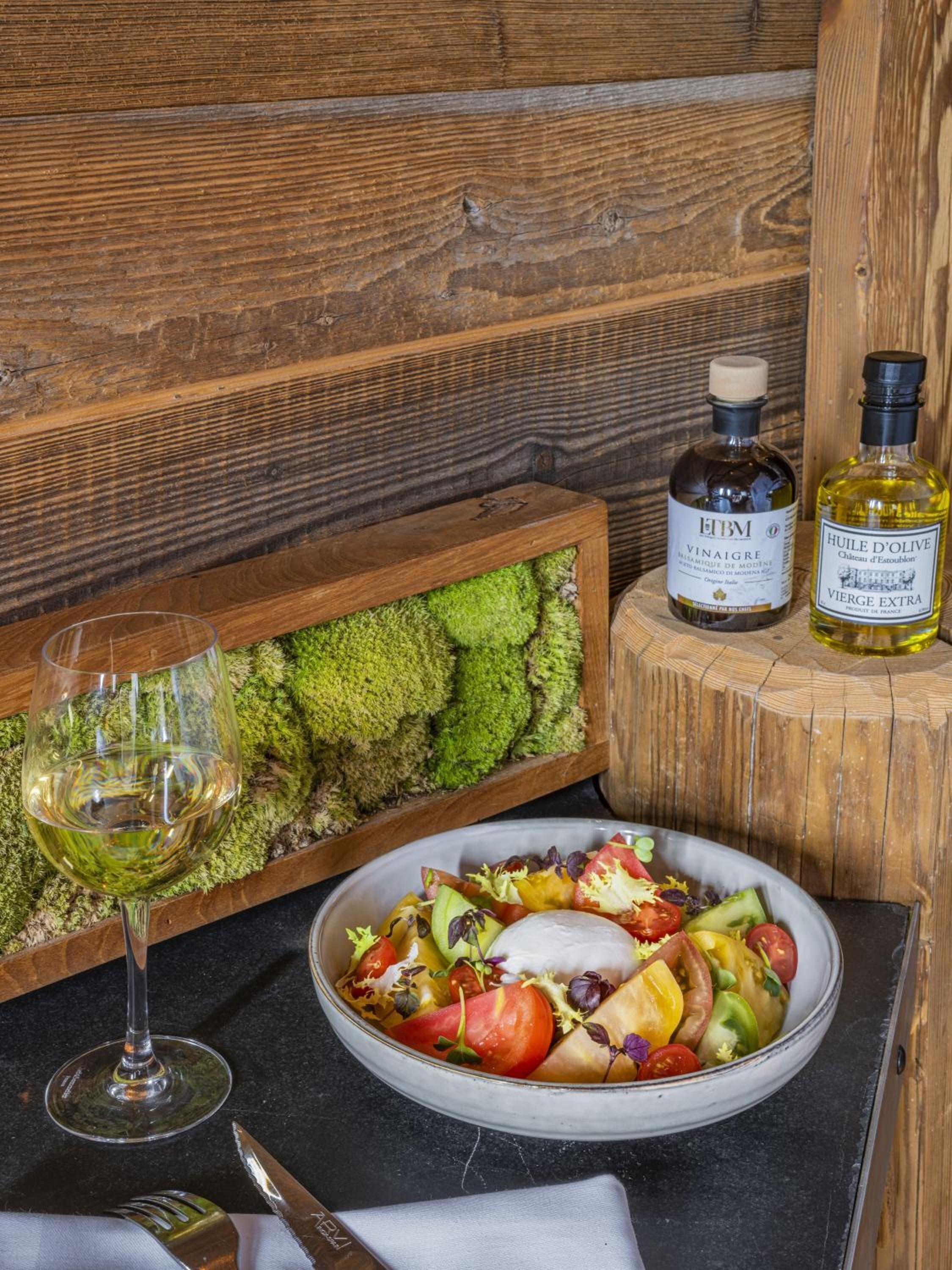 Une table en bois avec un verre de vin blanc, une bouteille de vinaigre EtBM et une bouteille d’huile d’olive.