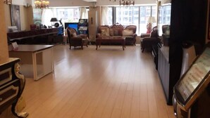 Un amplio salón con un sofá grande, una mesa de centro y un televisor. Hay una zona de cocina con mostrador y refrigerador. La habitación tiene ventanas grandes y una araña.
