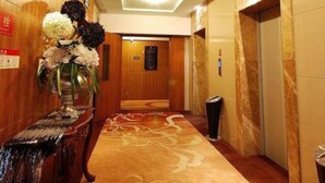 Un pasillo de hotel con un mostrador de recepción de madera, un jarrón de flores, un ascensor y suelo alfombrado.