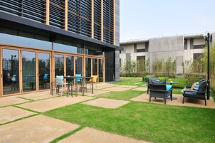 A modern outdoor patio with a seating area, a glass-enclosed building, and a garden with bamboo plants.