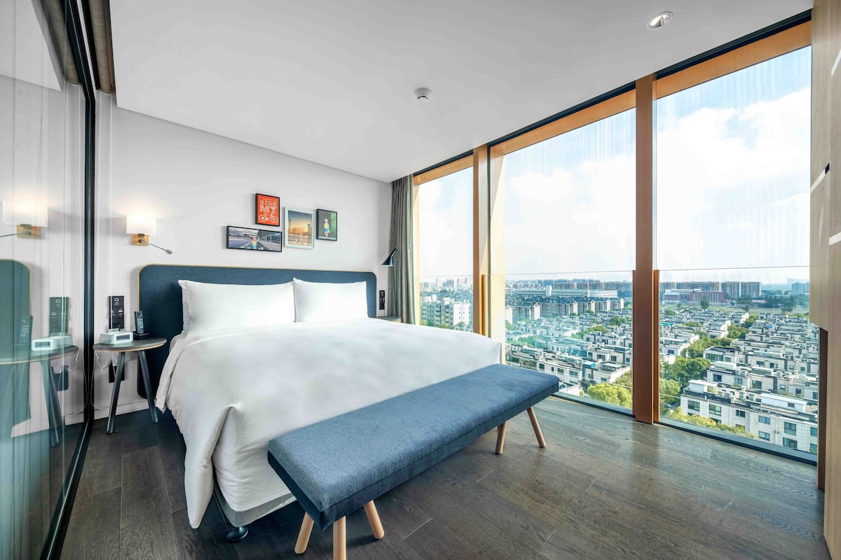 Une chambre d’hôtel moderne dotée d’un grand lit, d’une banquette, d’une table de chevet, d’une petite table et offrant une vue sur la ville grâce à de grandes fenêtres.