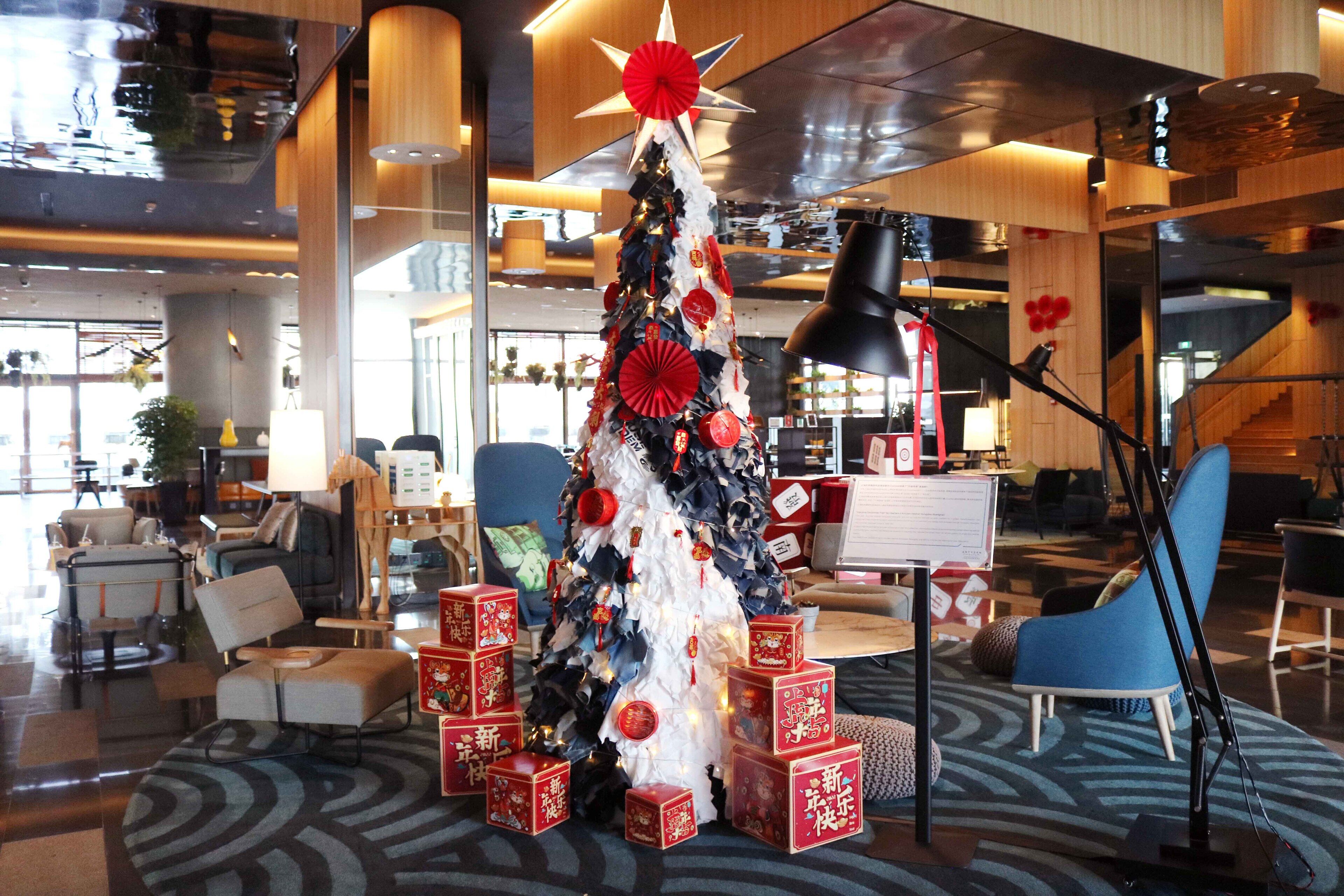 Eine moderne Hotelhalle mit einem Weihnachtsbaum, geschmückt mit roten Ornamenten und einem Stern auf der Spitze, umgeben von Geschenkkartons.