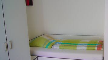 Una habitación pequeña con cama individual, paredes blancas, suelo de madera, un armario blanco y una manta a rayas de colores.