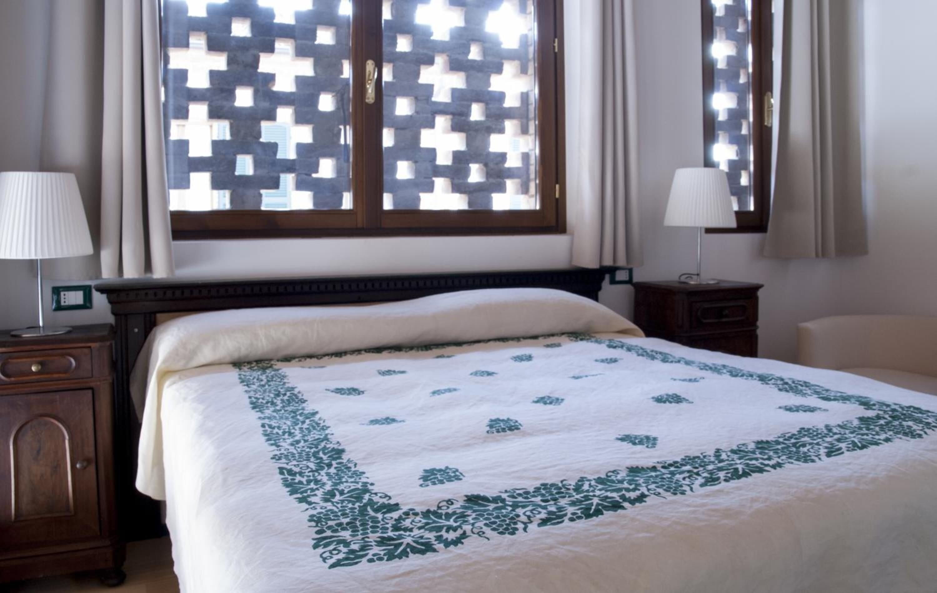 Una cama bien hecha con una colcha blanca y verde estampada, un cabecero de madera y una ventana con diseño de celosía.