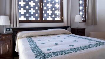 Una cama bien hecha con una colcha blanca y verde estampada, un cabecero de madera y una ventana con diseño de celosía.