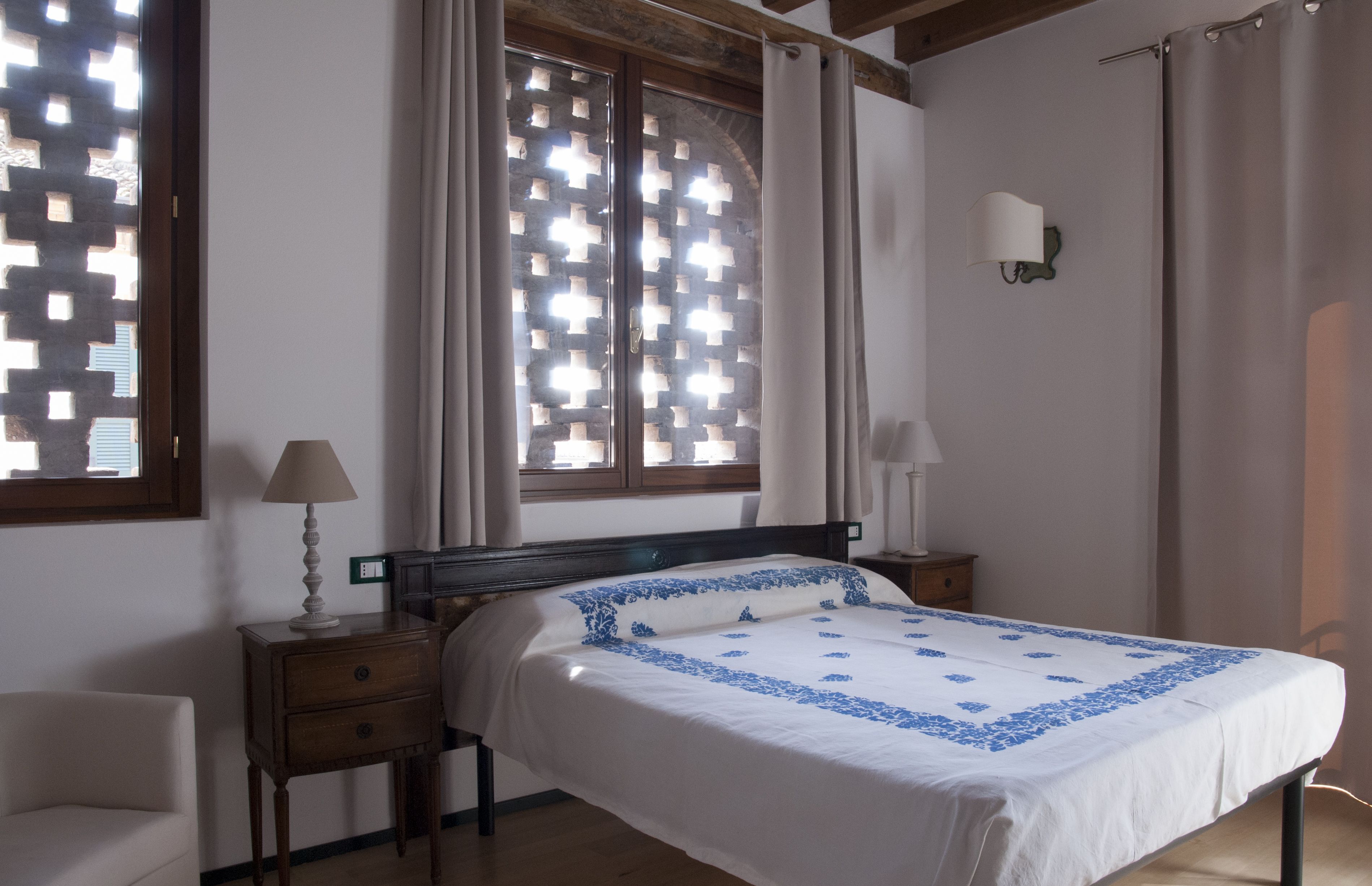 Un dormitori amb un llit, dues taules de nit, una petita taula auxiliar, una làmpada de paret i una finestra amb una pantalla decorativa.