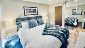 Una habitación de hotel con una cama, lámparas de noche, un cuadro en la pared, una puerta y un espejo.