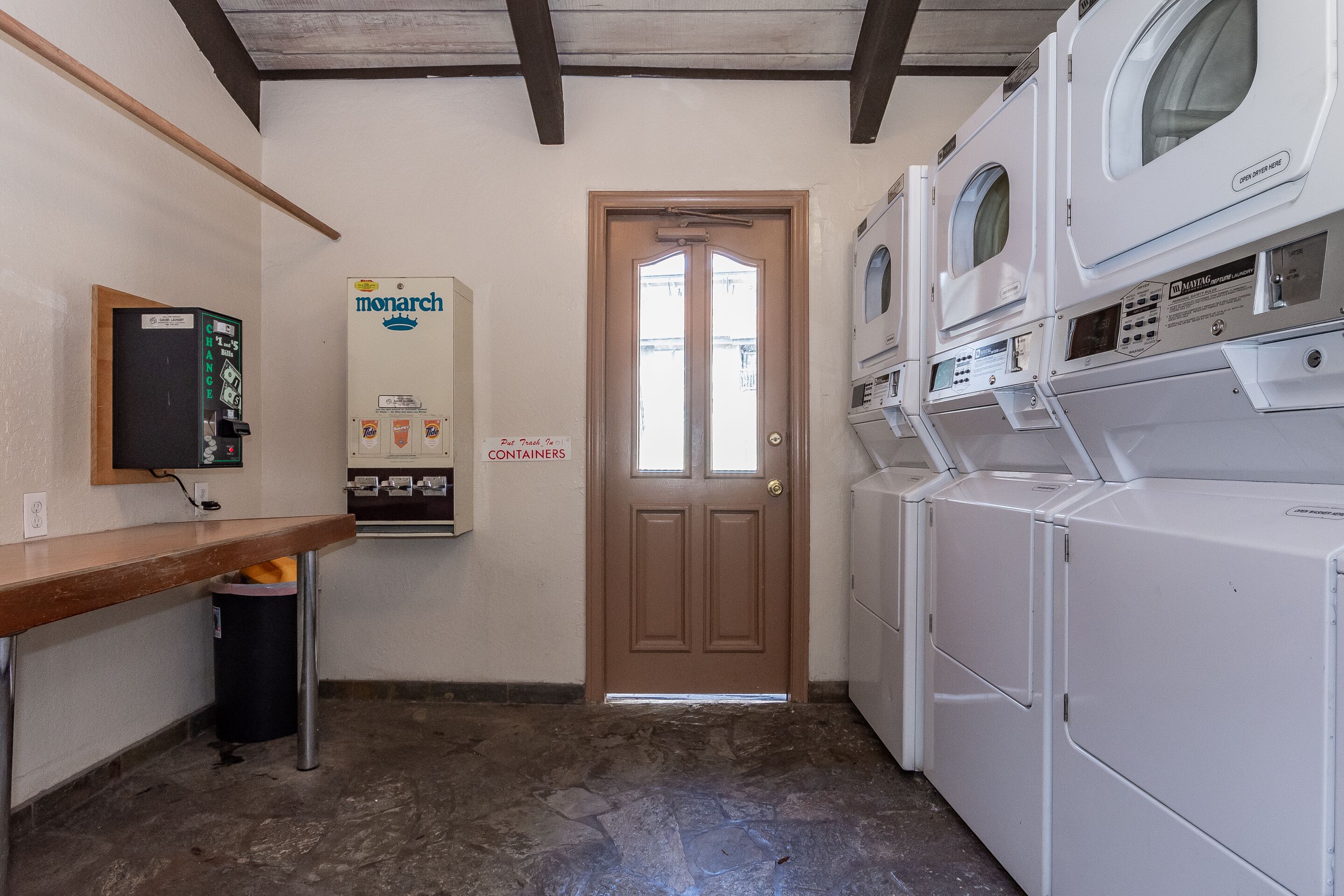 Une buanderie équipée d’une machine à laver et d’un sèche-linge superposés, d’un banc en bois, d’une poubelle et d’un distributeur de lessive Monarch.