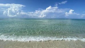 A beach with clear water, a sandy shore, and a blue sky with clouds.