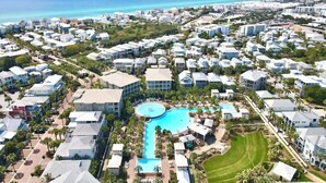 An aerial view of a coastal resort with a central swimming pool, multiple buildings, and a clear view of the ocean.