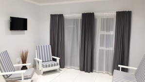 A modern living room with a white sofa, striped armchairs, a flat-screen TV, and large windows with grey curtains.