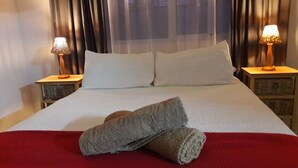 A neatly made bed with white linens and two pillows, a red blanket, and two rolled towels on the bed. There are two bedside tables with lamps on either side of the bed.