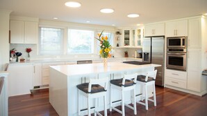A modern kitchen with white cabinetry, a central island with bar stools, stainless steel appliances, and a vase with flowers on the island. - Scenic Lake View Home (Tacoma)
