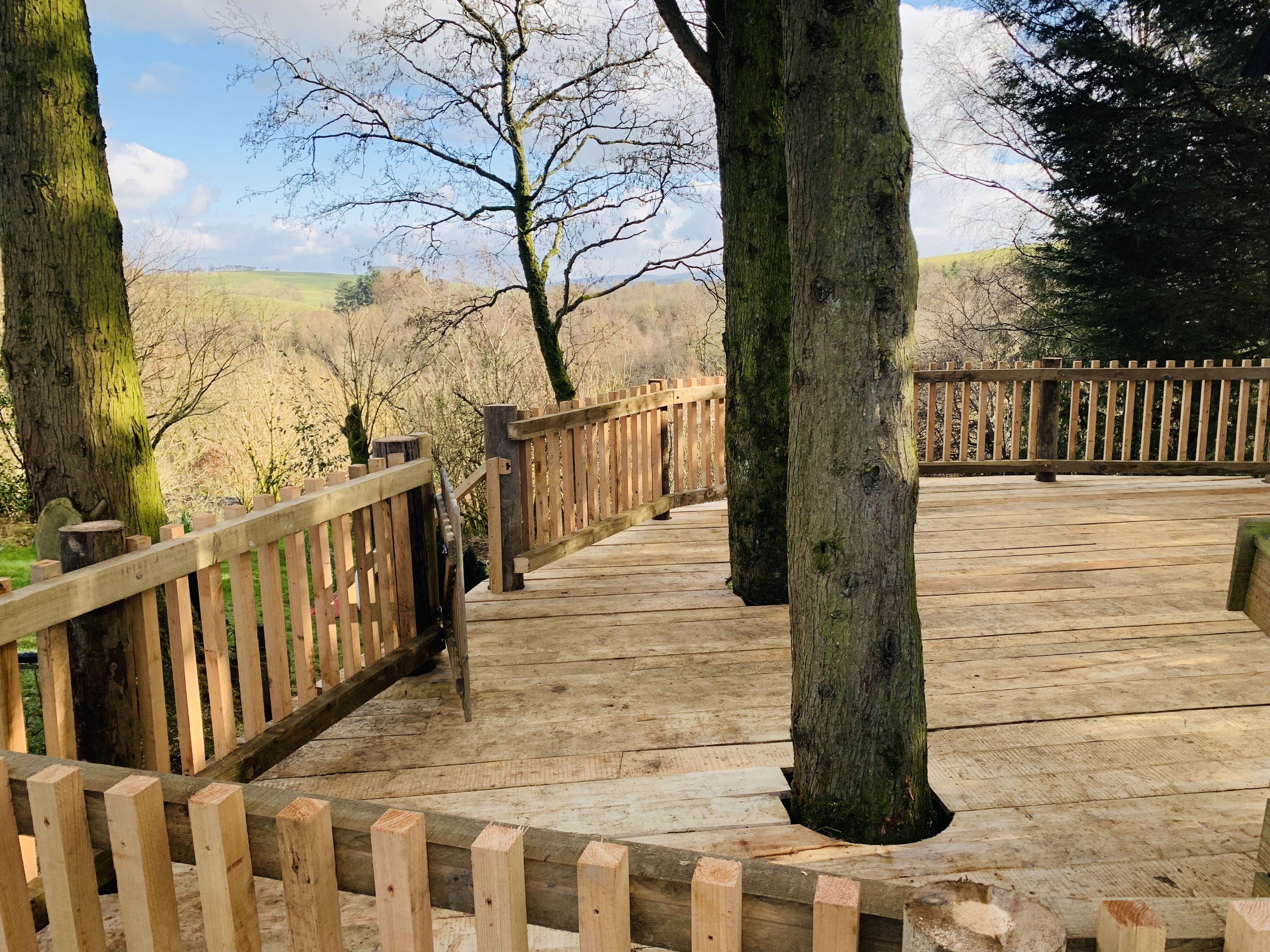 Una terraza de madera con barandilla, rodeada de árboles y vistas al campo.