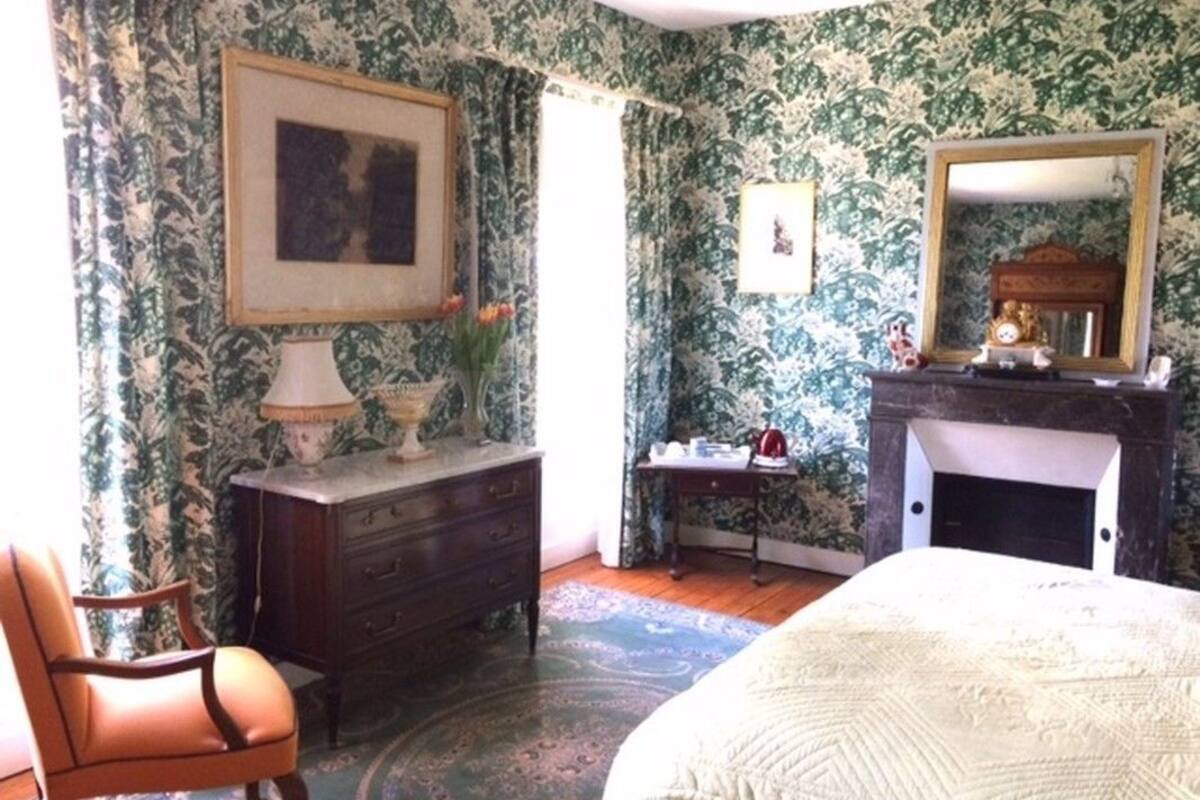Une chambre à coucher avec un papier peint à motifs floraux, un lit, une commode, une chaise, une cheminée et un miroir.