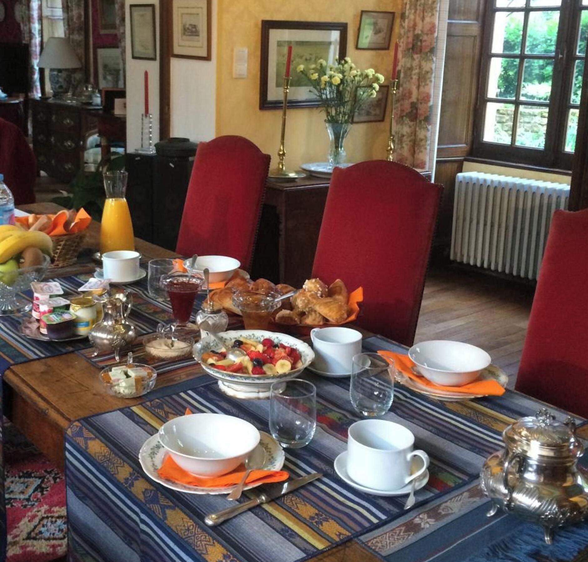 A dining table set with various dishes, including fruits and bread, in a room with red chairs and a view of greenery outside.