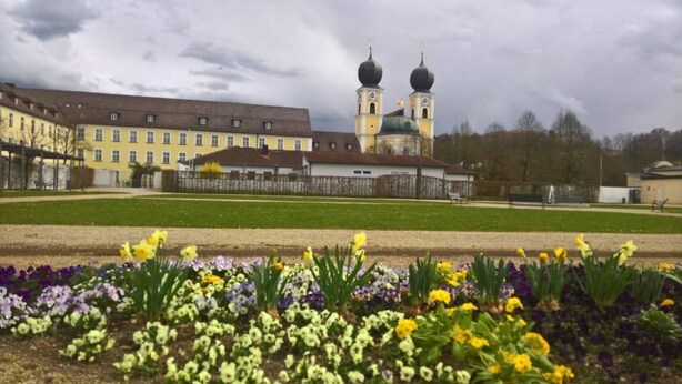A large building with a yellow facade and two domed towers, surrounded by a well-maintained lawn and flower beds. - Mettner Hof (Metten)
