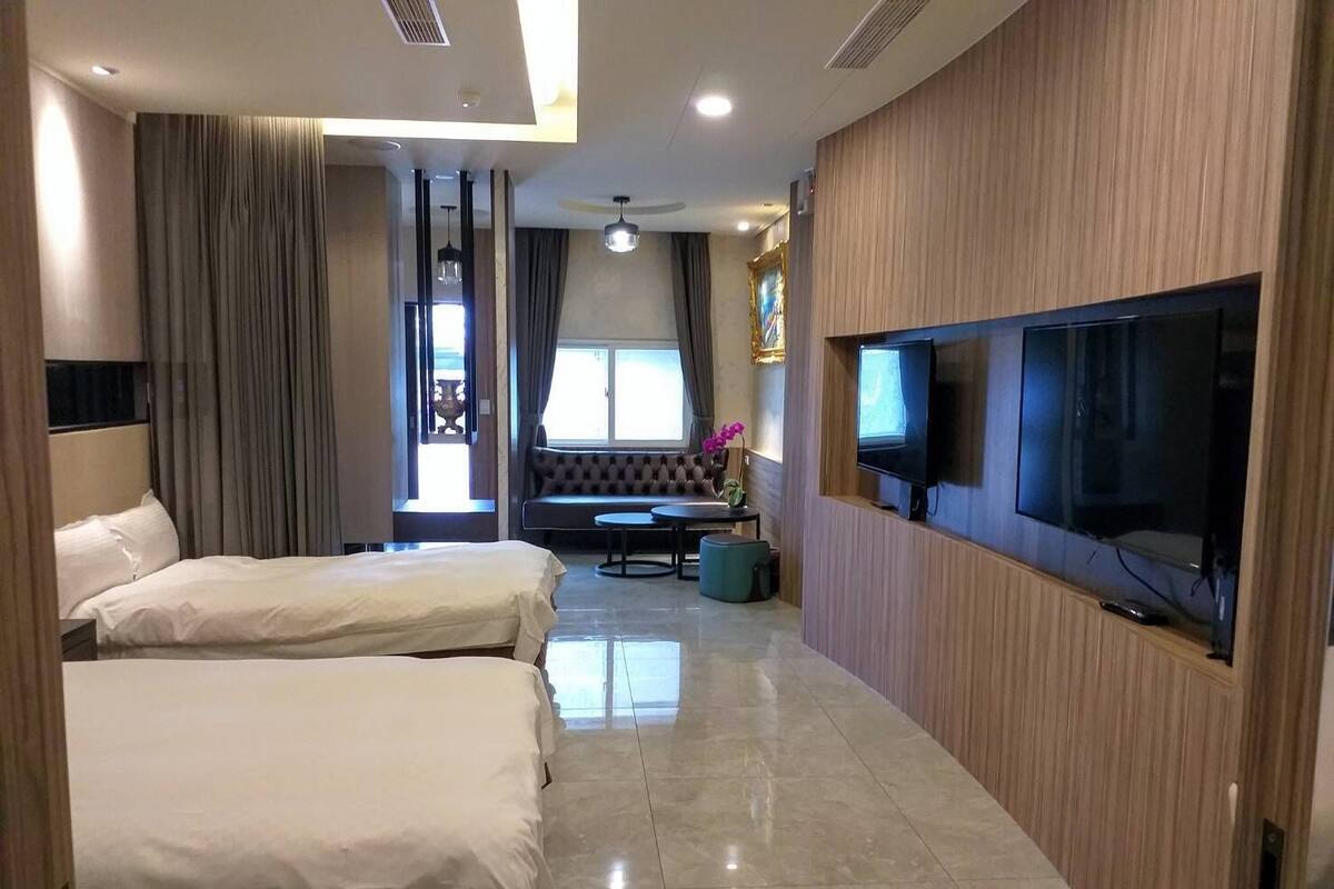 A hotel room with two beds, a TV mounted on the wall, a seating area with a sofa and a table, and a large window with curtains.