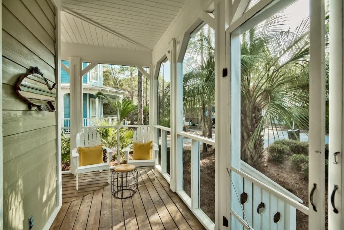 A covered porch with a wooden floor, white railings, and a view of a house and palm trees.