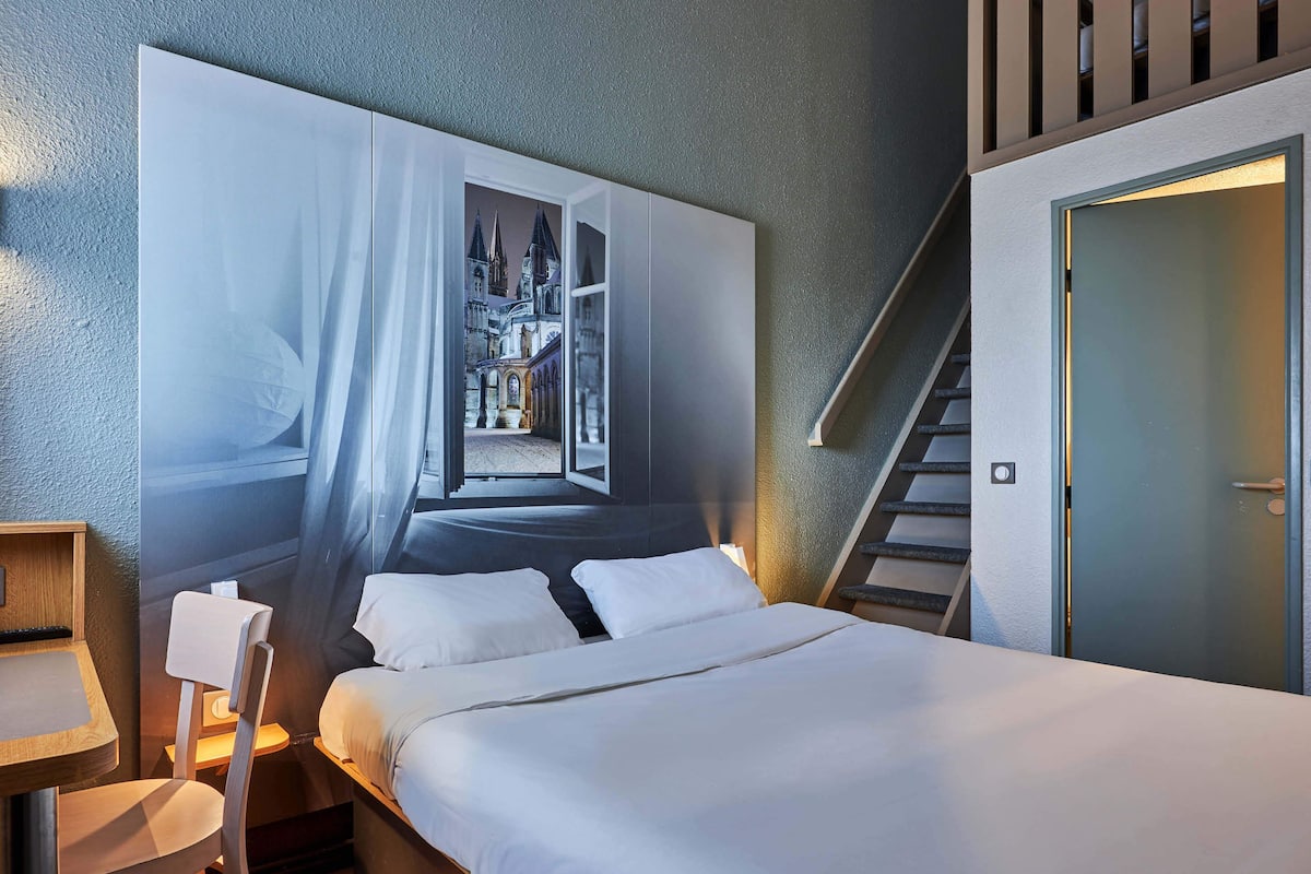 A hotel room with a large bed, a desk, a chair, and a wall-mounted artwork depicting a castle.
