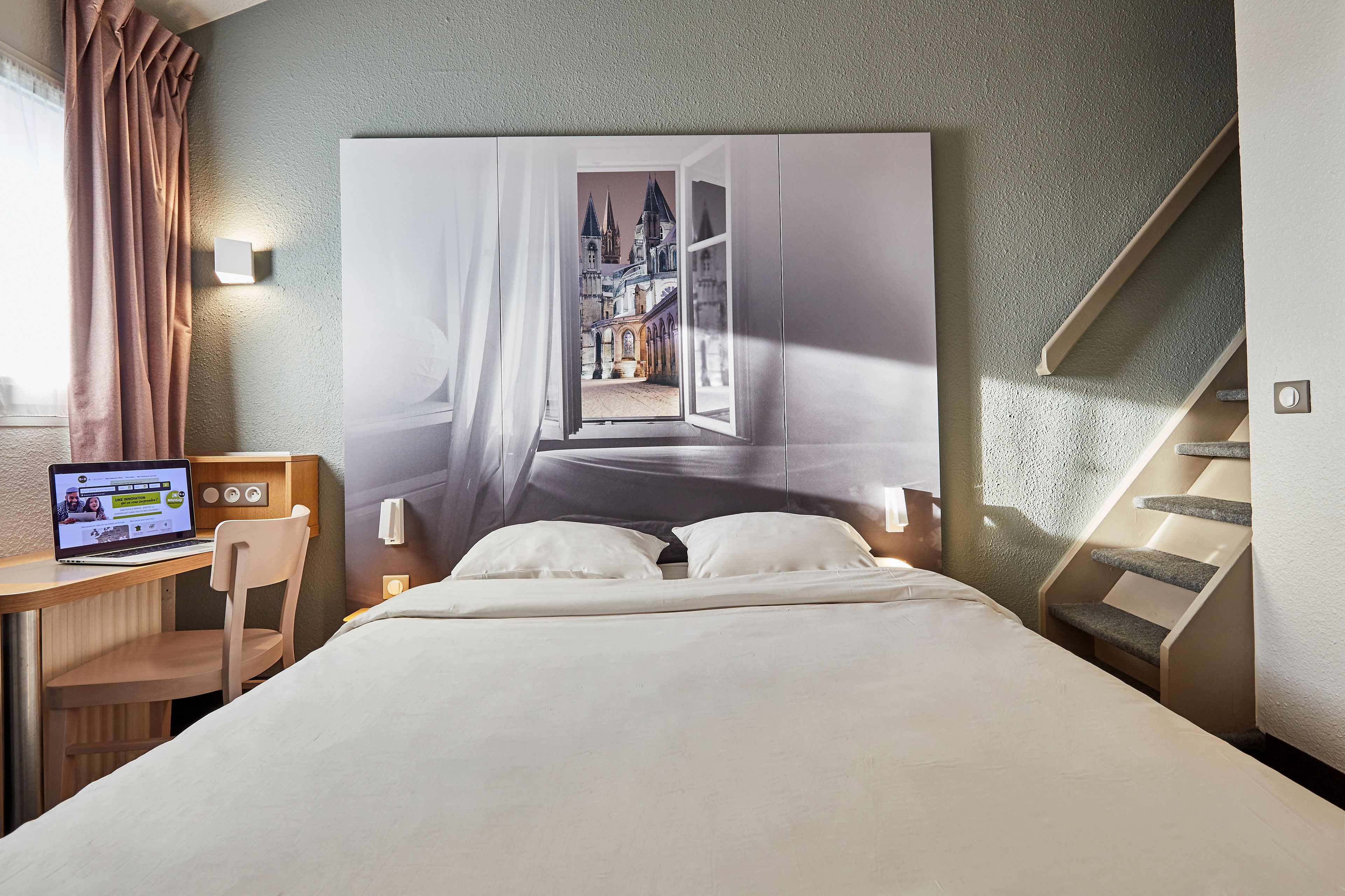 Habitación de hotel con cama, escritorio con una computadora portátil, silla y vista a un castillo desde una ventana abierta.