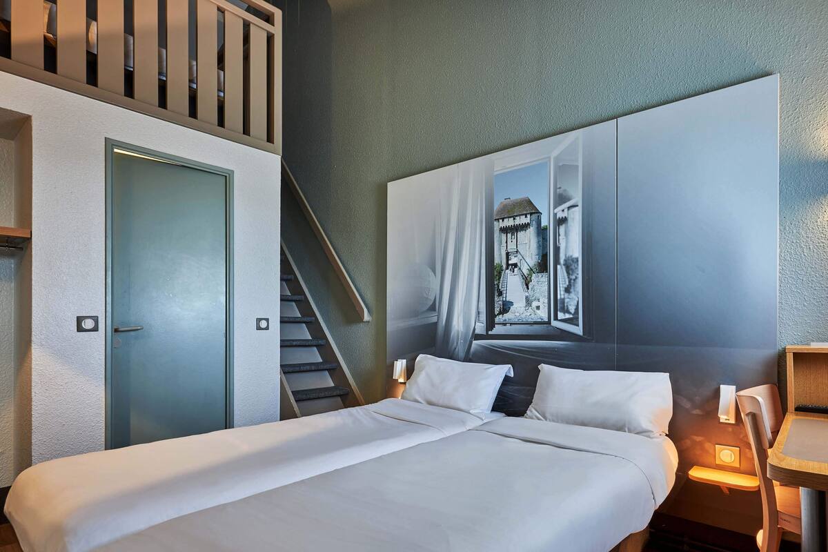 A hotel room with a bed, a nightstand, a chair, a desk, a window, and a staircase.