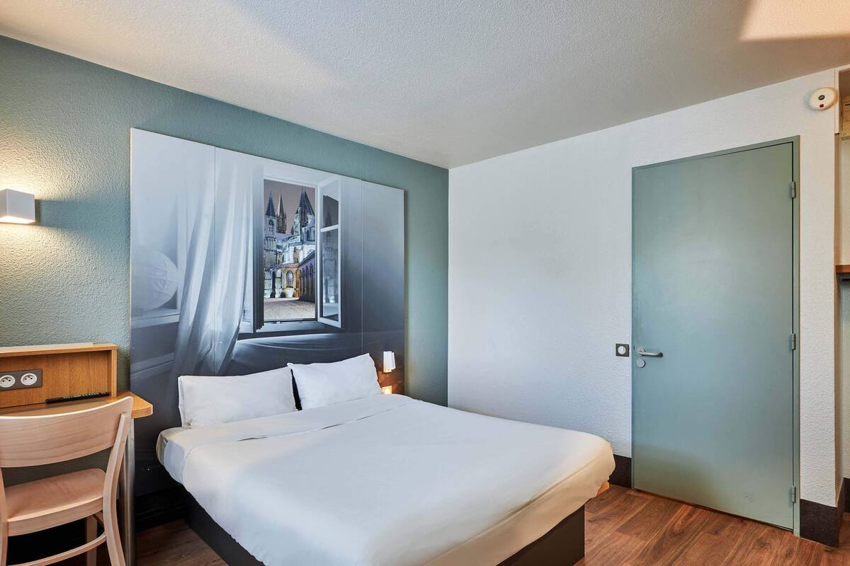 A hotel room with a bed, a desk, a chair, a large wall mural, a door, and a light switch.
