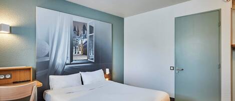 A hotel room with a bed, a desk, a chair, a large wall mural, a door, and a light switch.