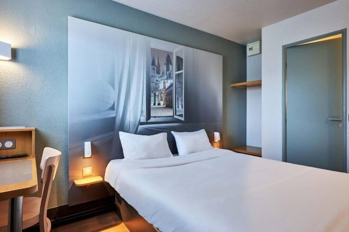 A hotel room with a bed, a desk, a chair, a nightstand, and a wall-mounted shelf.