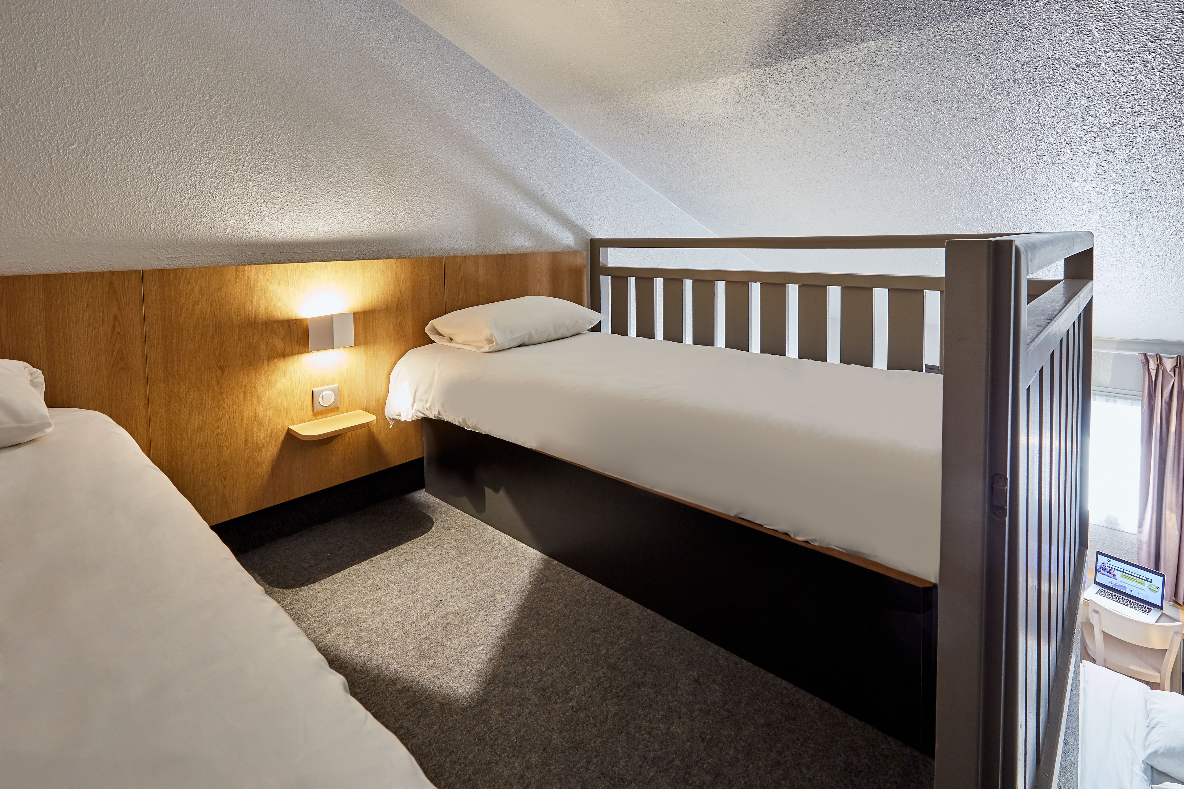 A small, well-lit room with a single bed, a bedside table with a lamp, and a laptop on a small desk.