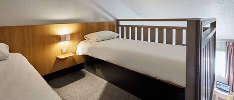 Una habitación pequeña y luminosa, con una cama individual, una mesita de noche con lámpara y un ordenador portátil sobre un escritorio pequeño.