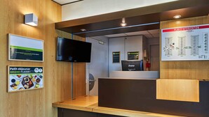 A reception area with a wooden reception desk, a flat-screen TV, and a 'Plan d'intervention' poster on the wall.