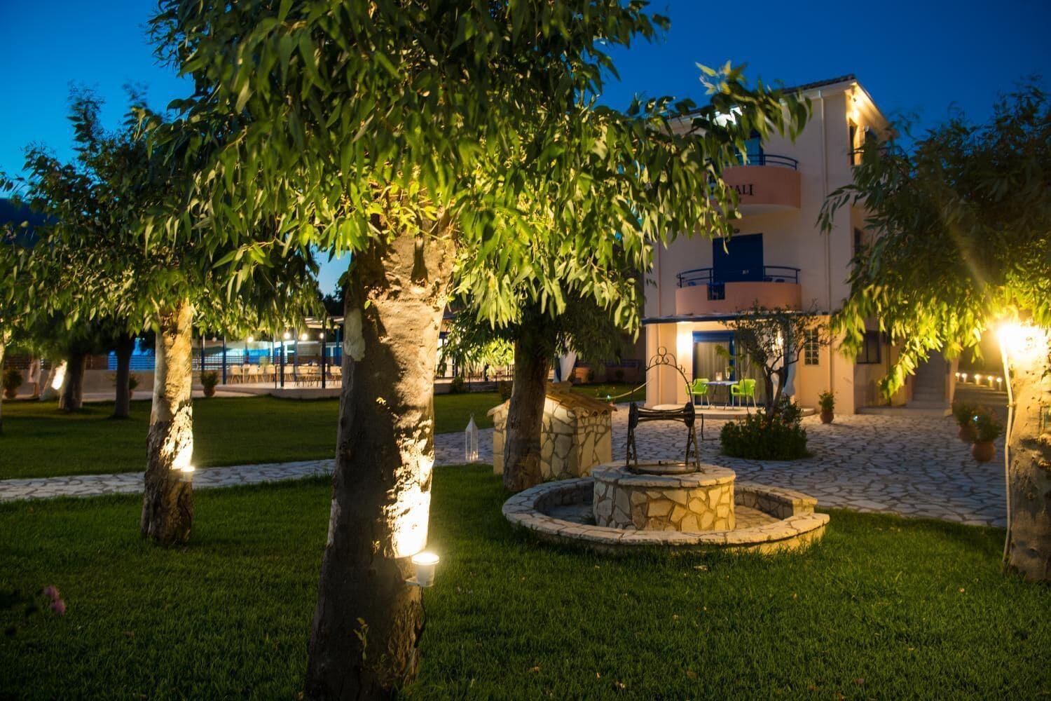 A well-lit outdoor area with a stone fountain, trees, and a building with a balcony.