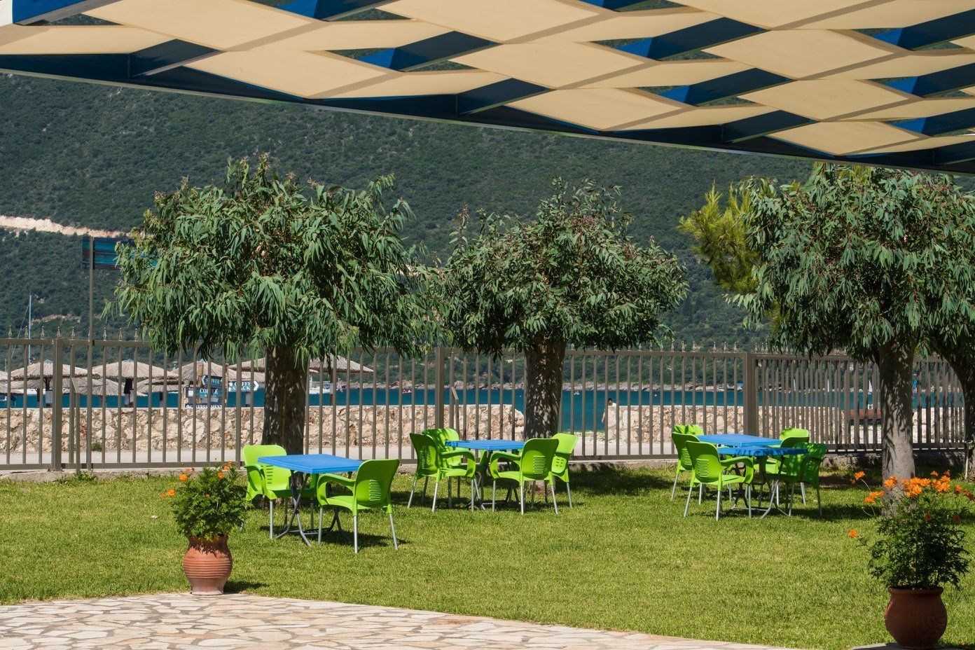 Een terras met groene stoelen en blauwe tafels, omringd door bomen en uitzicht op een wateroppervlak.