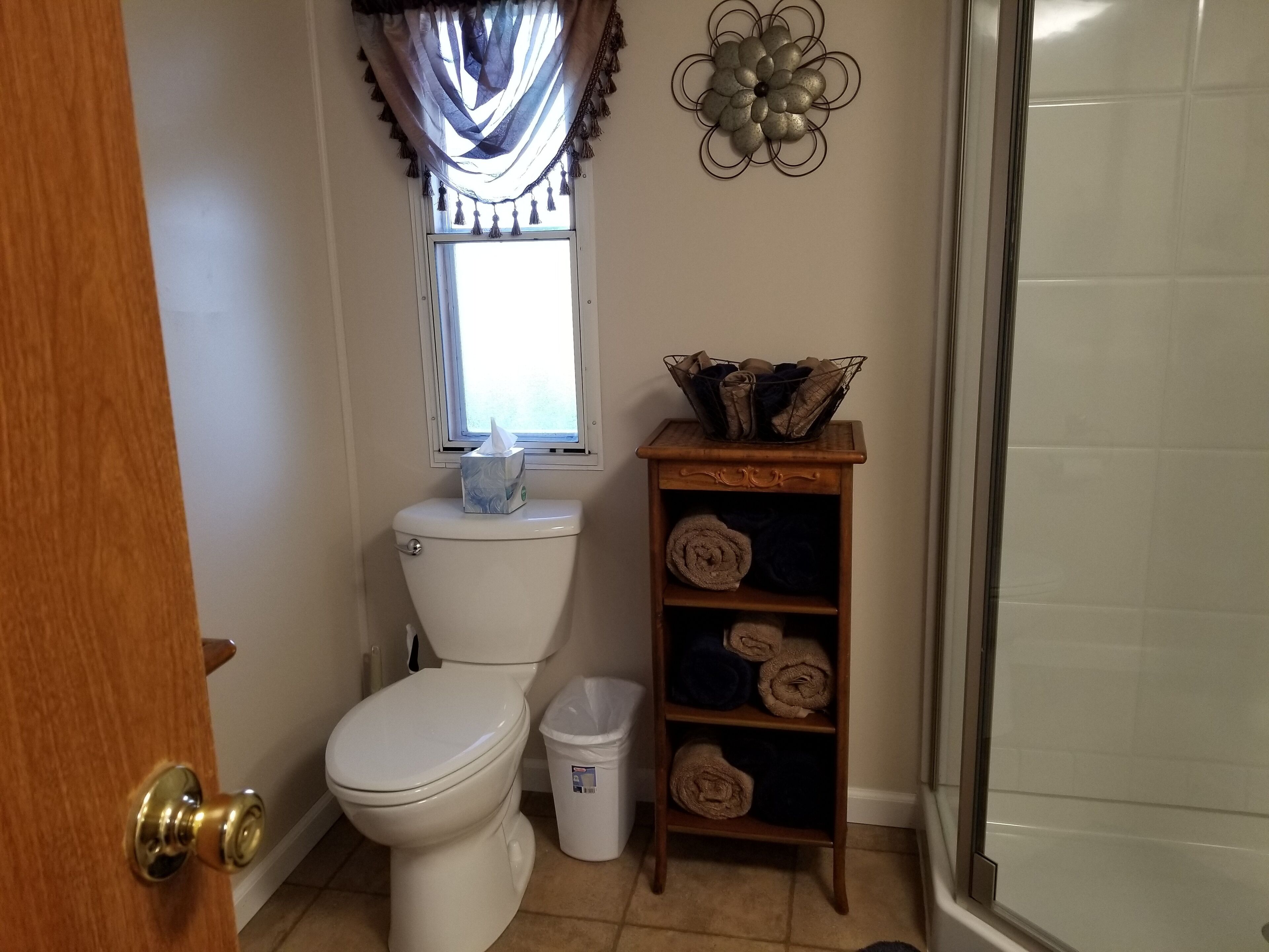Une salle de bain comprenant des toilettes, une fenêtre avec des rideaux, un élément décoratif en métal, un porte-serviettes avec des serviettes et une poubelle.