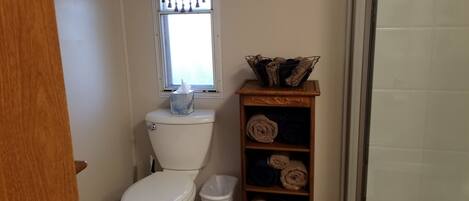 Une salle de bain comprenant des toilettes, une fenêtre avec des rideaux, un élément décoratif en métal, un porte-serviettes avec des serviettes et une poubelle.